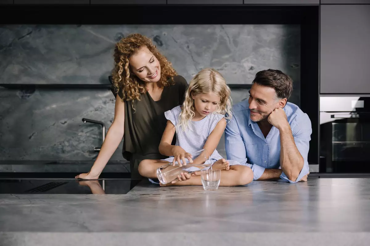 Mann und Frau stehen hinter Küchenblock, Kind sitzt auf Arbeitsfläche und schenkt Wasser in ein Glas ein